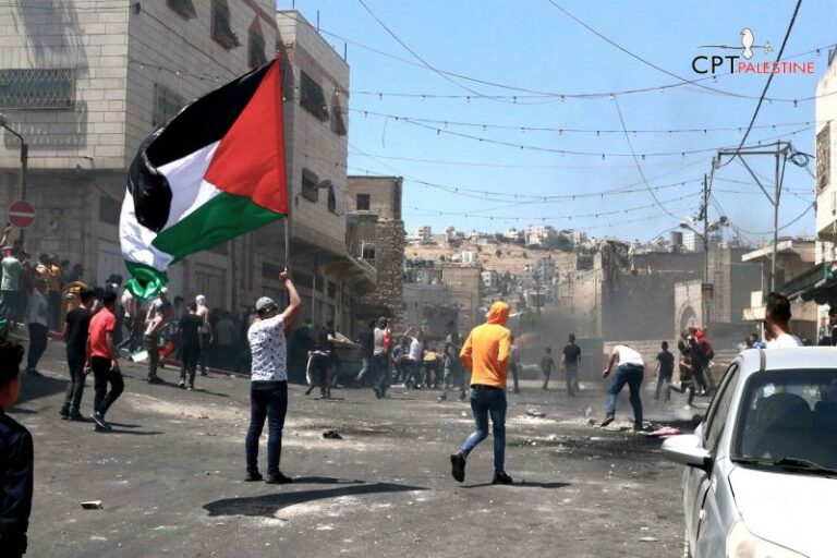 Dozens of young people are in the street that is filled with smoke and tear gas. One person on the left holds a very large Palestinian flag. There are white stone buildings on the right, and festive lights strung above the street. There is a white car on the left in the foreground.