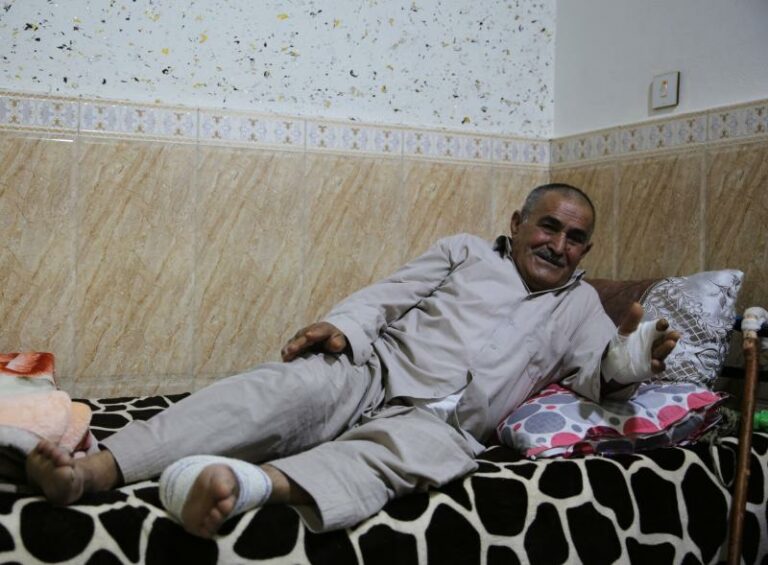 Ramazan Ali is laying on a bed, resting from injuries sustained from Turkish military bombing. Ramazan has bandages on his left foot and his left hand from the injuries. Ramazan’s left hand is on a pillow, and the bed has a black and white blanket. Ramazan is wearing a grey dress shirt and pants. Behind Ramazan is a brown and white wall.