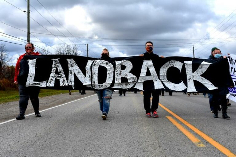 Cuatro personas caminan hacia la cámara en un camino, la línea amarilla divide a la tercera y cuarta persona. Llevan una pancarta negra con letras blancas en negrita que dice LANDBACK