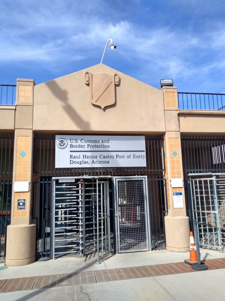 Una gran estructura de puerta se encuentra en el centro de la imagen, con un torniquete de metal. El letrero sobre el torniquete dice Servicio de Aduanas y Protección Fronteriza de EE. UU. Raul Hector Castro Port of Entry Douglas, Arizona