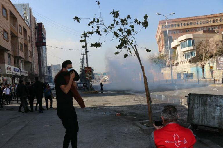 Hundreds of people gather in the street, many wearing backpacks. Tear gas plumes in the distance.