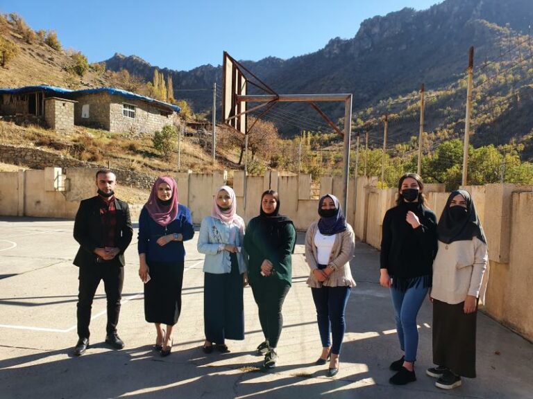 Siete profesores posan para una foto en el patio de hormigón de la escuela. Hay una red de baloncesto detrás de ellos, y en la distancia se puede ver una casa en la ladera de la montaña.