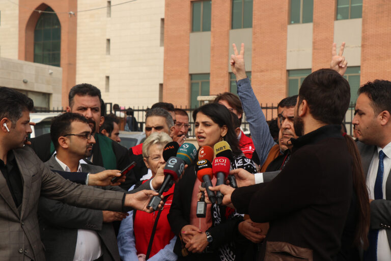 A woman surrounded by press microphones speaks at a press conference surrounded by journalists and supporters