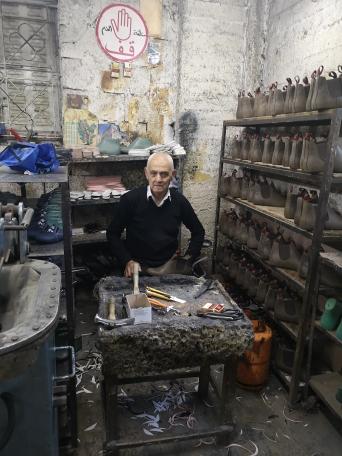 Abu Ashraf sits at his work table where he makes shoes. There are shoe molds lined up on shelves to his right