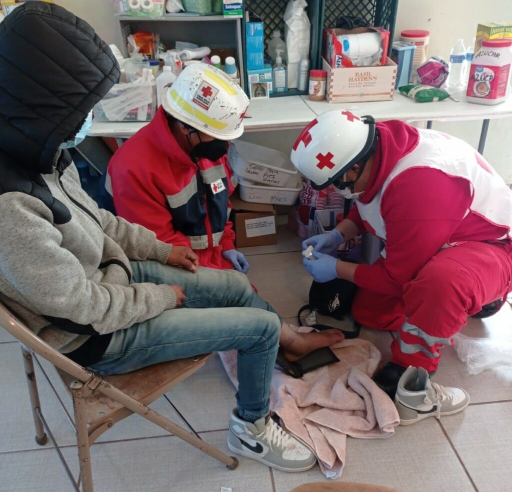 a person wearing a black hoodie and jeans sits on a chair while two Red Cross medics attend to their injured foot.