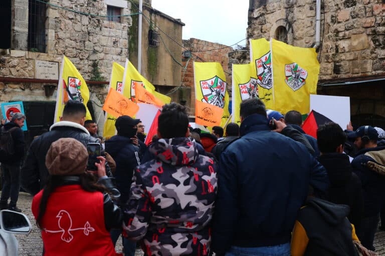 A CPT member stands to the left, wearing a toque and a red CPT vest, holding a camera aimed towards a crowd of people holding yellow flags and marching.