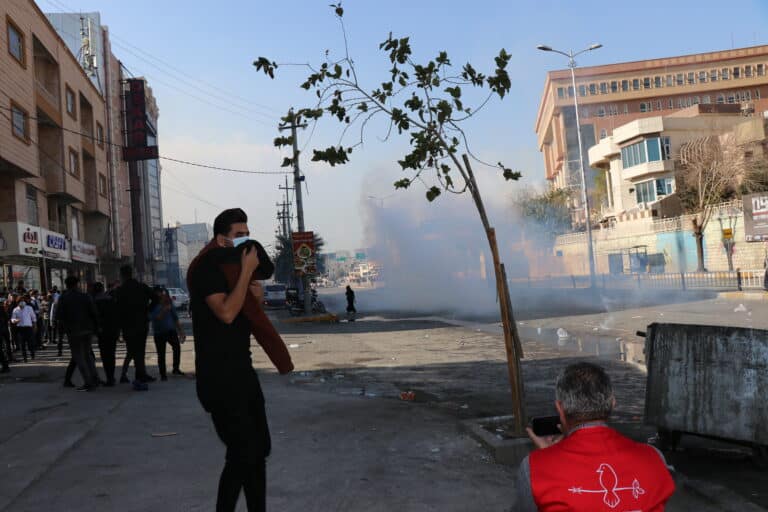 a person stands on the side of a street, wearing a surgical mask and covering their face with a piece of clothing. There is a large cloud of tear gas in the street. A person wearing a CPT vest crouches in the foreground, taking footage on a cell phone.