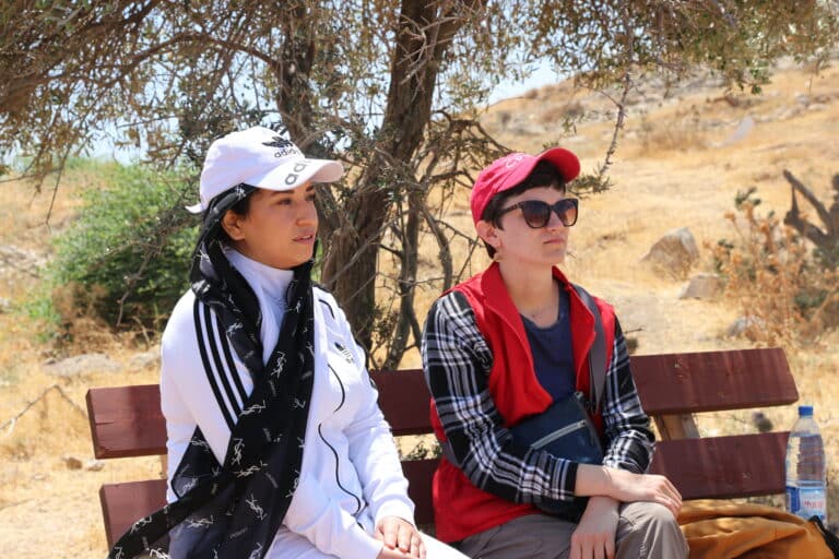 Two people sit on a bench, one wears a white baseball cap and jacket, and a black hijab. The other wears a red CPT cap and vest, and sunglasses.