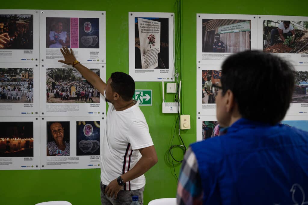 A man points to images on a green wall as a person in a blue jacket watches