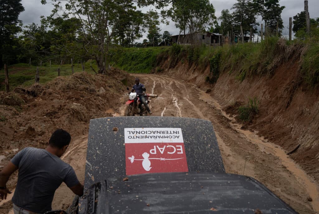 A vehicle with a bonnet open and a man standing beside it talking to a man on a motorcycle. The road is muddy with high embankments.