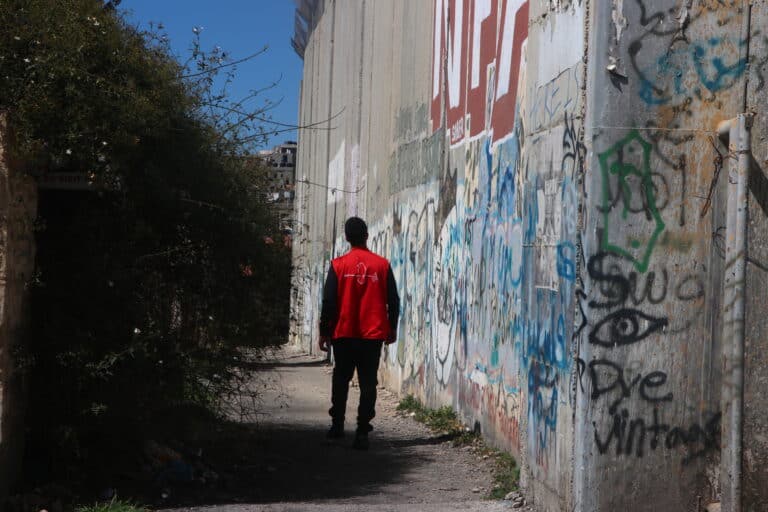 A person wearing a red CPT vest walks along a road with the apartheid wall to their right, covered in graffiti and towering over them.