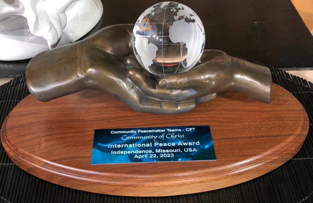 The sculpture award given to CPT shows two hands, the smaller hand cradled by the larger and and cradling a globe. Plaque reads Community Peacemaker Teams - CPT Community of Christ International Peace Award Independence, Missouri, USA April 22, 2023