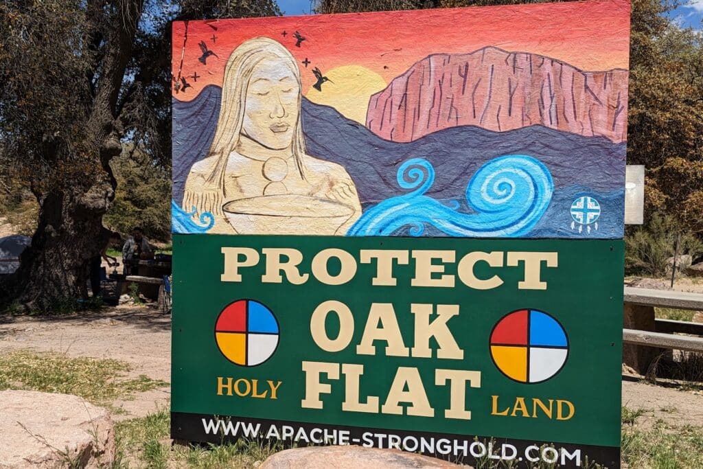 A sign reads "Protect Oak Flat Holy Land" with two medicine wheels and a painting of a person with air swirling around them, a large rock formation in the distance and a sunset.