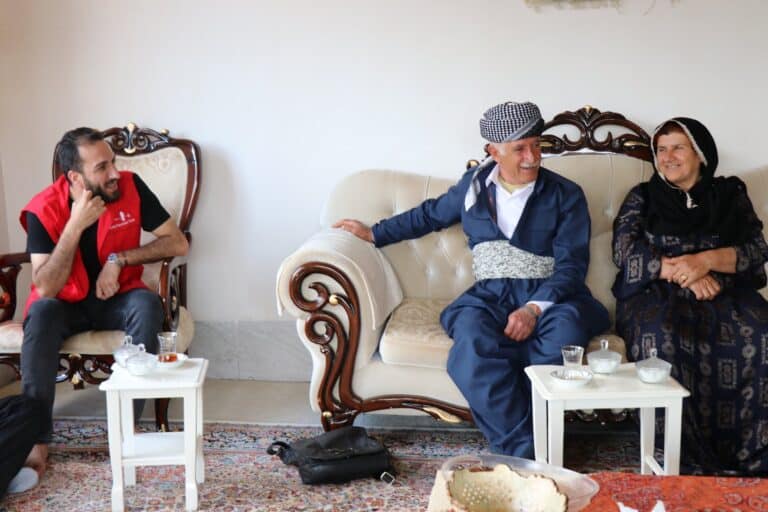 CPT member Kamaran Osman sits on a chair to the left, wearing a red CPT vest, and looking over at Kak Bapir and Aisha Khan sitting on a couch. There are small tables in front of them with tea.