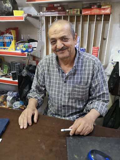 Musbah se sienta frente al cajero en su tienda, sonriendo a la cámara. Lleva una camisa a cuadros con cuello y hay estantes de productos detrás de él.