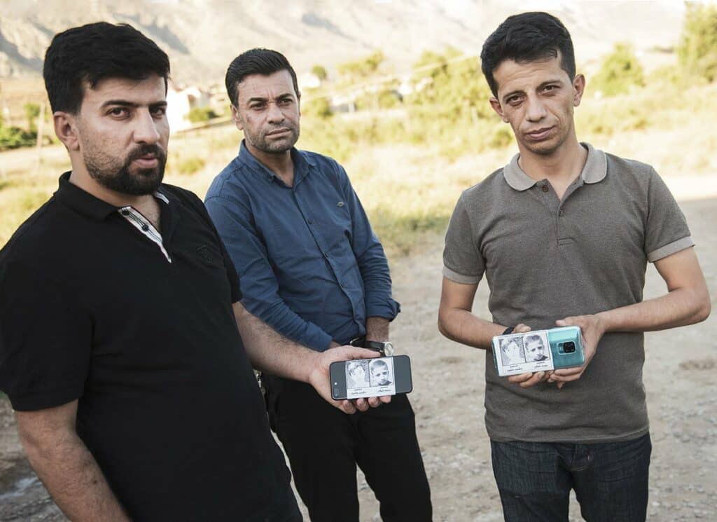 Three people stand facing the camera, two hold up phones with images of children who were killed. 