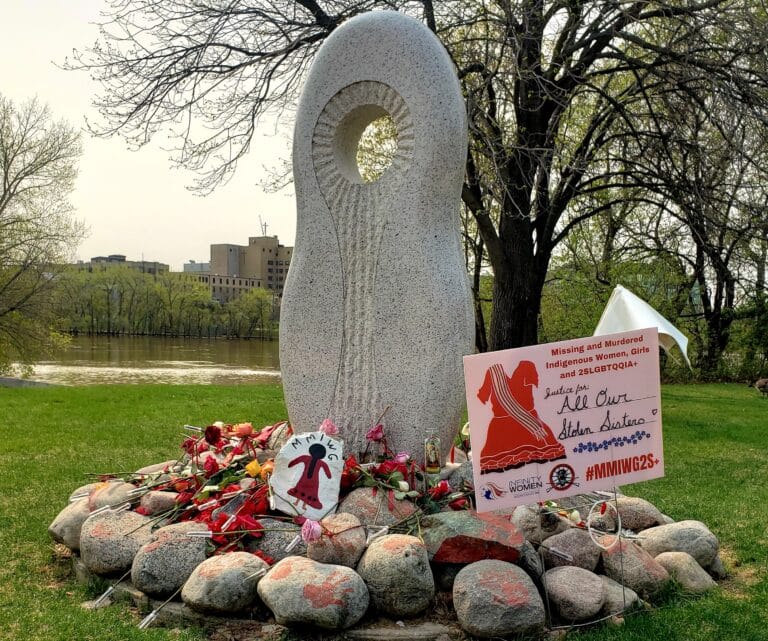 a stone monument stands, with several layers of stones laid at the base of the statue. A sign placed between the stones reads "Justice for all our sisters" #MMIWG2S+