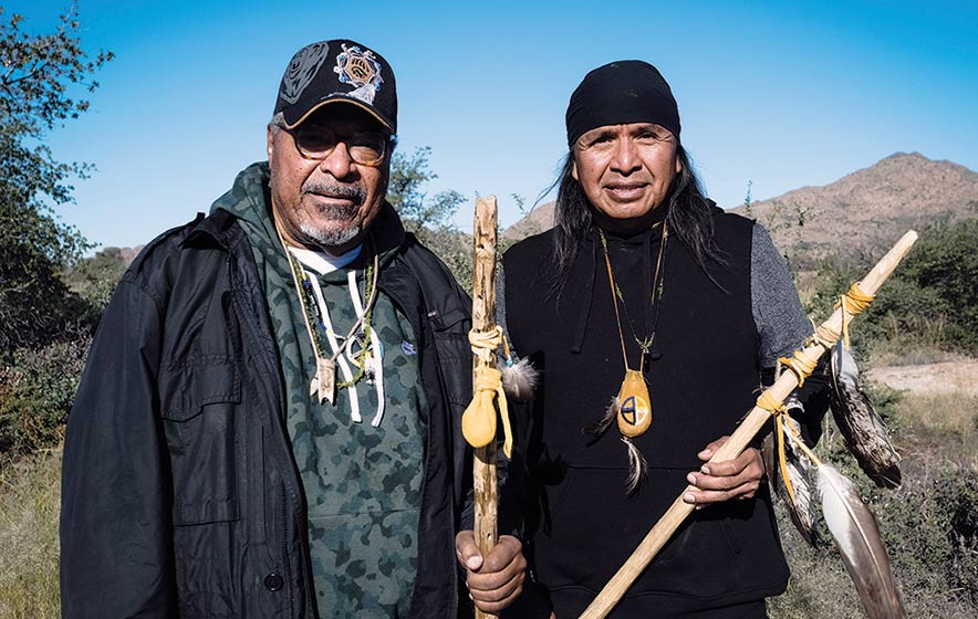 Dr. Wendsler Nosie and Rev. Dr. Mendez stand together, esch holding a staff and wearing necklace adornments.
