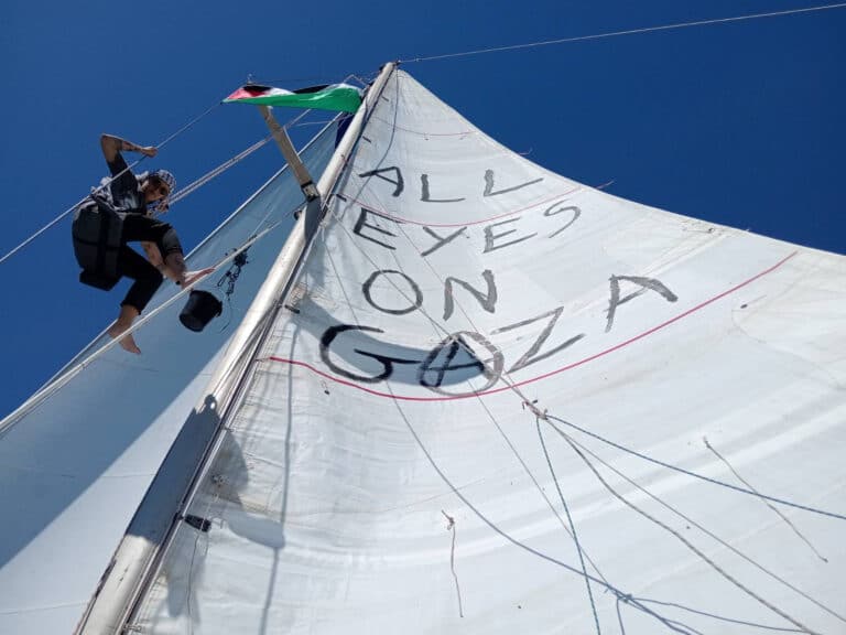 A boat crewmember next to a sail that reads "all eyes on Gaza"