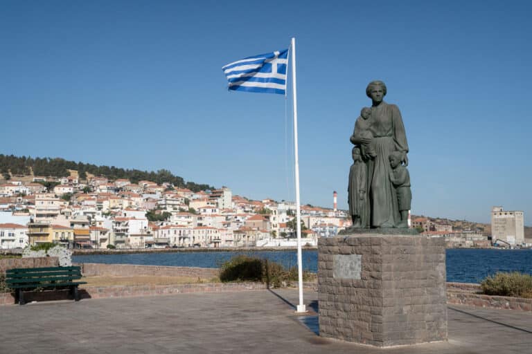 A statue of a migrant and her two children along side a waving flag of greece on the coastline of mytilene