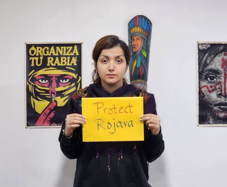 a woman holds a sign that reads protect rojava
