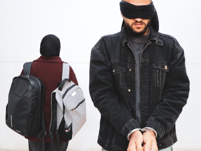 A man is handcuffed and blindfolded and a woman carries two backpacks