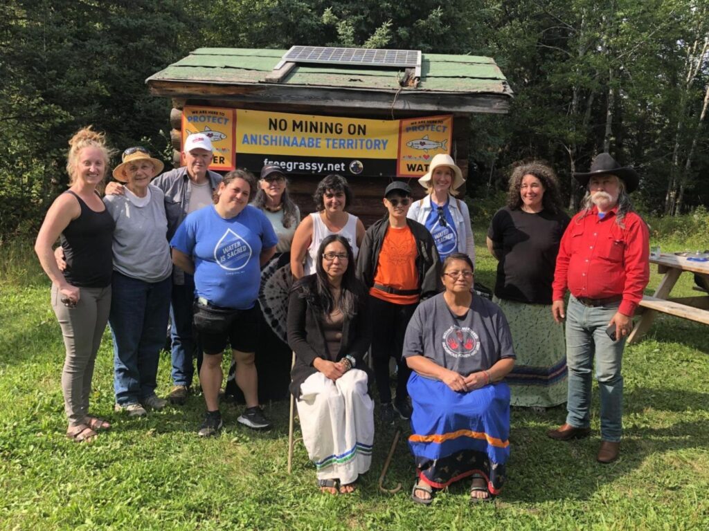 CPT Delegation to Turtle Island members with Grassy Narrows First Nations members. The sign in the background reads “No Mining on Anishnaabe Territory. freegrassy.net”