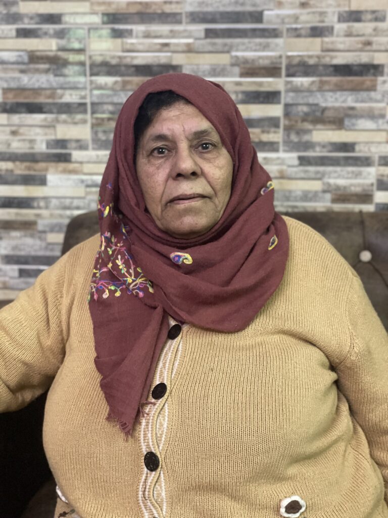 A woman wears a red hijab and a brown sweater, looking straight into the camera.