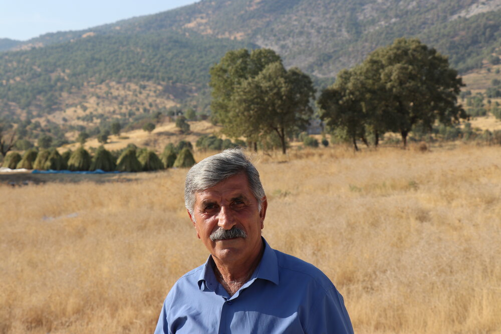 Kak Auetr stands on the road. His home lies behind the trees, right under the mountains covered in ash.