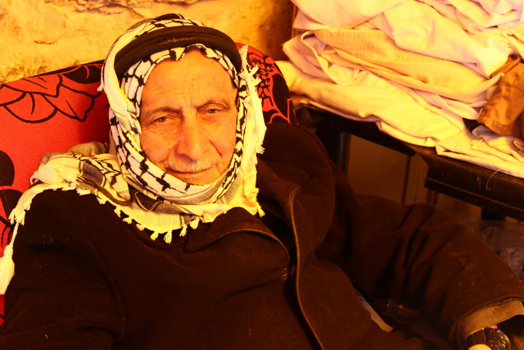 Mohammad wears a black and white Palestinian kuffiyeh on his head and is seated on a couch.