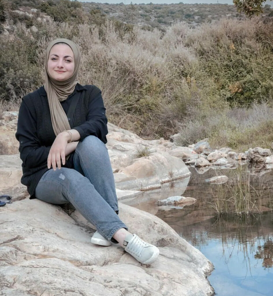 A woman sits on a rock beside a small body of water, there are dry bushes behind her. She wears jeans, a black top, and a beige hijab and smiles at the camera.